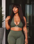 A woman modeling an olive green two-piece activewear set consisting of a scoop-neck crop top and matching high-waisted capri leggings, holding a yellow handbag.