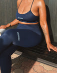A woman wearing a navy blue Schuix activewear set, consisting of a sports bra and matching leggings, sitting on a black bench against a dark wood-paneled wall.