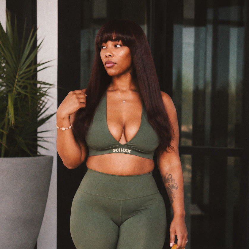 A woman modeling an olive green two-piece activewear set consisting of a scoop-neck crop top and matching high-waisted capri leggings, holding a yellow handbag.