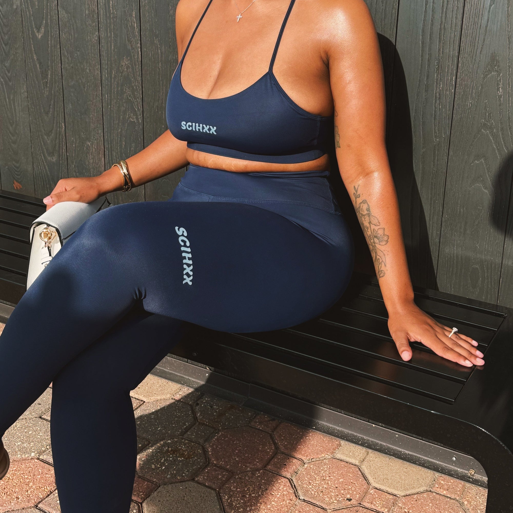A woman wearing a navy blue Schuix activewear set, consisting of a sports bra and matching leggings, sitting on a black bench against a dark wood-paneled wall.