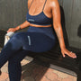 A woman wearing a navy blue Schuix activewear set, consisting of a sports bra and matching leggings, sitting on a black bench against a dark wood-paneled wall.
